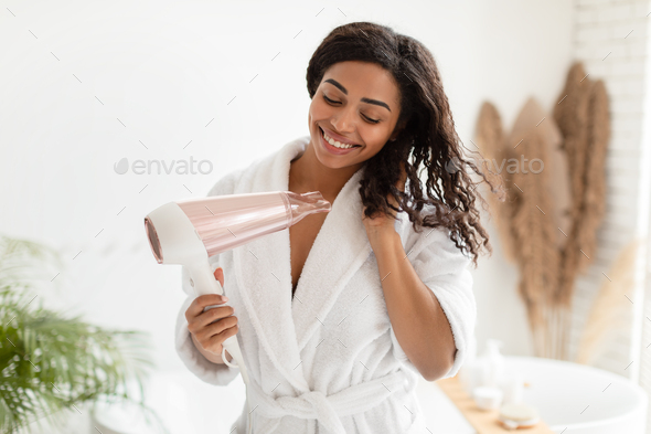 Happy Black Lady Drying Wavy Hair With Hairdryer In Bathroom