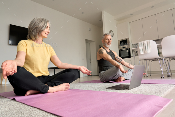 Happy mature older couple doing yoga meditating together at home with laptop.