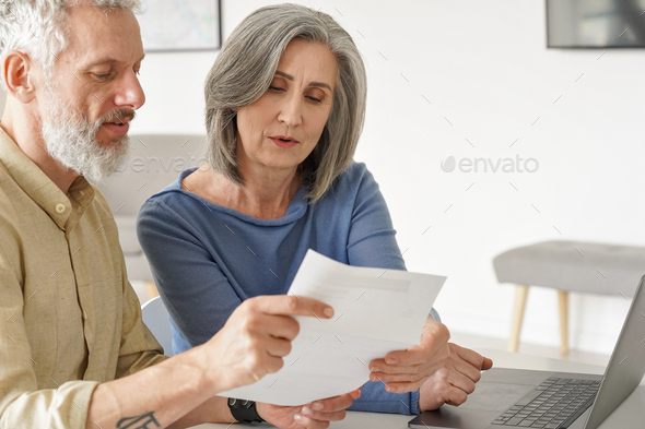 Older mature couple checking bank documents or bills using laptop at home.