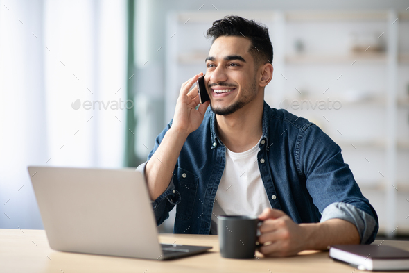 Happy arab guy freelancer working from home, having phone conversation
