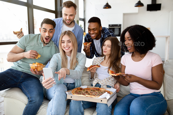 Positive diverse friends having home party, sitting on couch with pizza, chips and smartphone