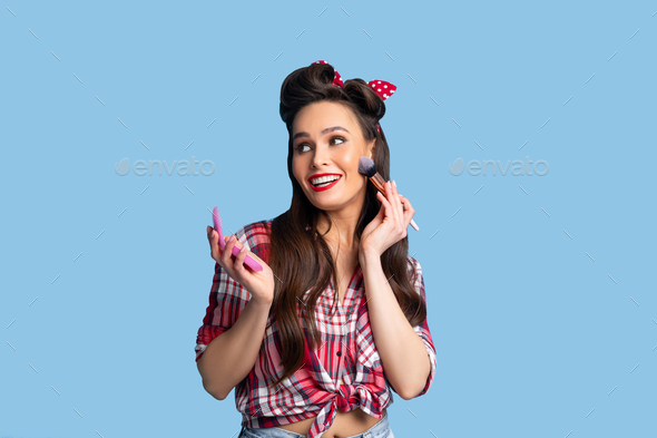 Bright makeup. Sexy young pinup lady in retro outfit putting on decorative cosmetics with brush over