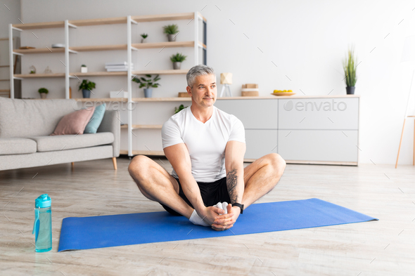 Active mature man sitting in butterfly asana , doing yoga on sports mat at  home in living room Stock Photo by Prostock-studio