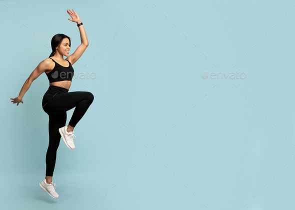 Fitness concept. Fit young african american woman jumping, lifting leg up, blue background with