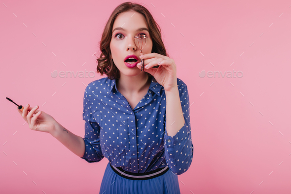 Shocked white girl in trendy blouse curls her eyelashes on pink background. Studio portrait of spec