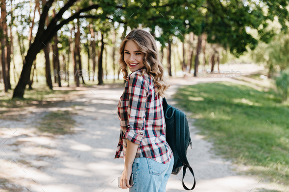 Glad girl in checkered shirt and blue jeans standing in park. Inspired  woman with leather backpack Stock Photo by look_studio