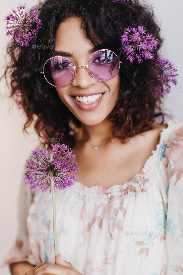 Excited brown-eyed african girl posing with flowers at home. Indoor portrait of sensual black lady