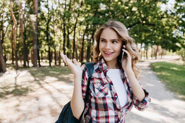 Pretty girl in good mood calling friend while walking in park. Outdoor shot of pleasant curly woman