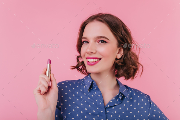 Laughing caucasian woman with dark hair holding pink lipstick. Indoor shot of ecstatic girl in good