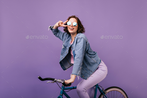 Pleasant stylish woman posing on purple background on bike. Indoor photo of smiling ecstatic girl s