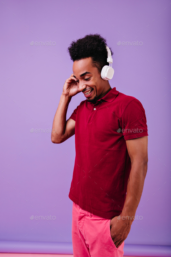 Amazing guy with wavy hair looking down while listening music. Indoor photo of pleasant african mal