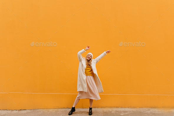 Cheerful, bright blonde dances with raised arms on orange background in white knitted clothes