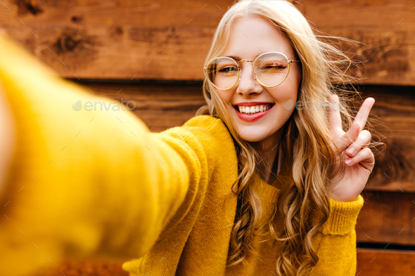 Close-up self portrait of a curly blonde in a bright sweater. Happy young lady makes peace sign and
