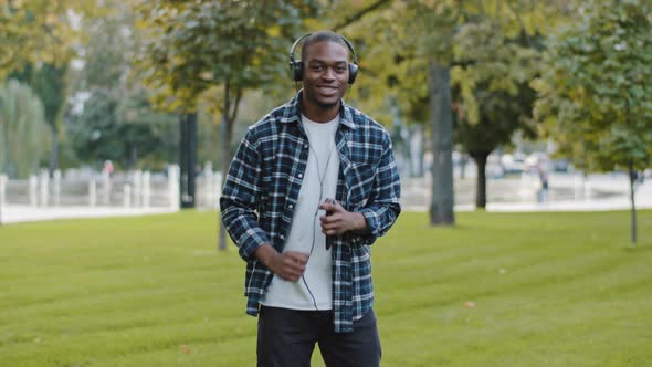 African American Stylish Cheerful Carefree Man Young Happy Guy in Park Wears Headphones Uses Mobile alt