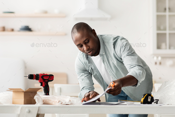 Concentrated african american man reading furniture installation instruction, assembling kitchen