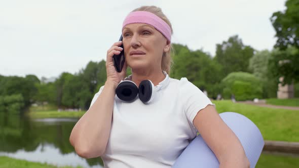 Happy Smiling Senior Woman with Exercise Mat Talking on Smartphone at Park alt