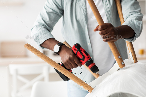 Furniture assembly process. African american handyman screwing legs to chair with electric