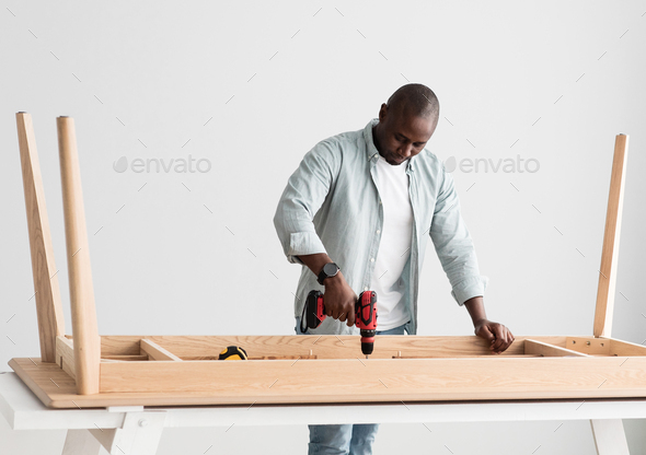 Professional african american craftsman drilling wooden planks of table, assembling new furniture