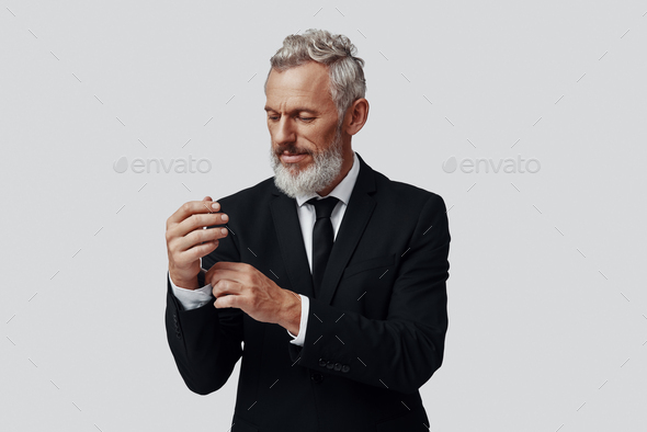 Elegant mature man in full suit adjusting sleeve while standing against grey background