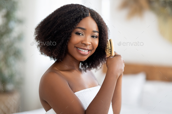 Portrait of happy young black lady brushing her curly dark hair at home
