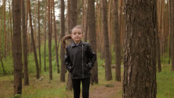The Little Girl Runs Along the Path Road in the Pine Forest. Dressed in the Leather Jacket. alt