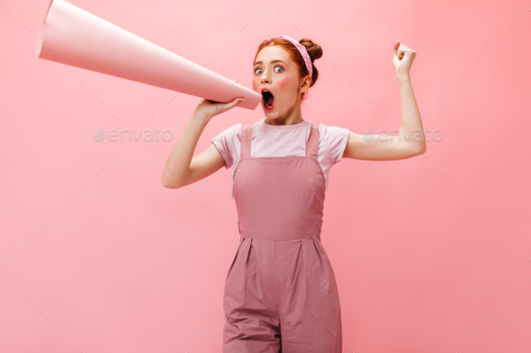 Joyful young lady in pink dress and white T-shirt screaming into mouthpiece on pink background