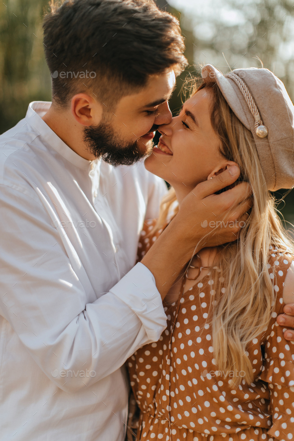 Lady in polka-dot silk dress and her man in white linen shirt with smile hugging and posing in park