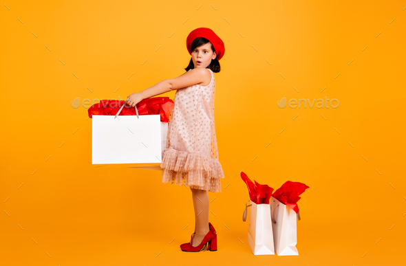 Amazed kid in red beret and mom\'s shoes standing on yellow background. Studio shot of cute child wi