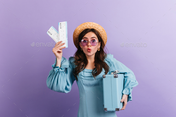 Funny woman in boater and lilac glasses stares into camera in amazement, showing her plane tickets