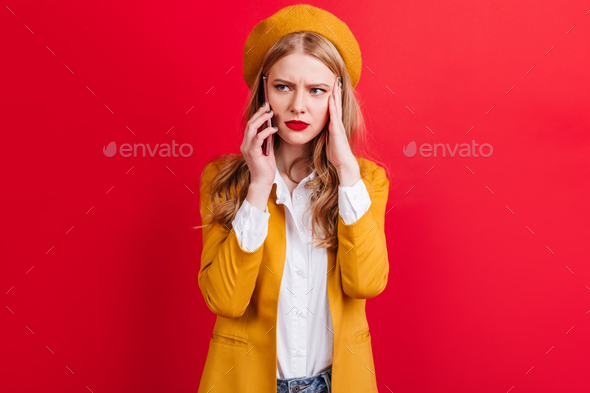 Irritated blonde woman in yellow beret talking on phone. Studio shot of caucasian girl in jacket ho - Stock Photo - Images