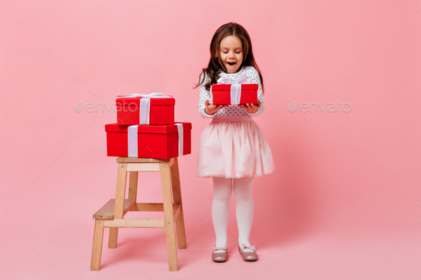 Little girl in white smart outfit looks happily at red box with gift, wanting to open it as soon as