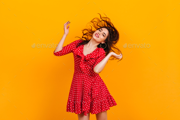 Stylish young girl in red short dress plays hair. Portrait of brunette in hoop earrings on yellow b