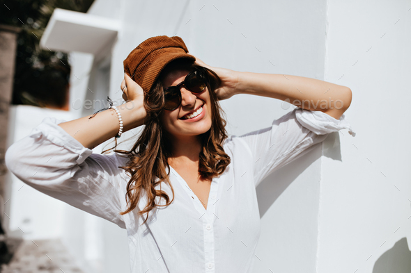 Beautiful woman in sunglasses is wearing corduroy cap. Portrait of girl in high spirits against whi