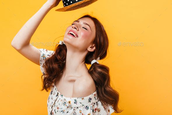 Young girl with rosy cheeks takes off her hat and laughs. Portrait of woman with ponytails on orang