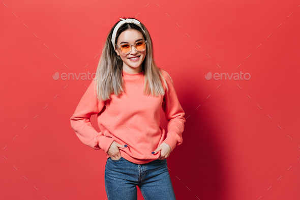 Girl with hair band, wearing orange glasses and sweatshirt, posing on red background