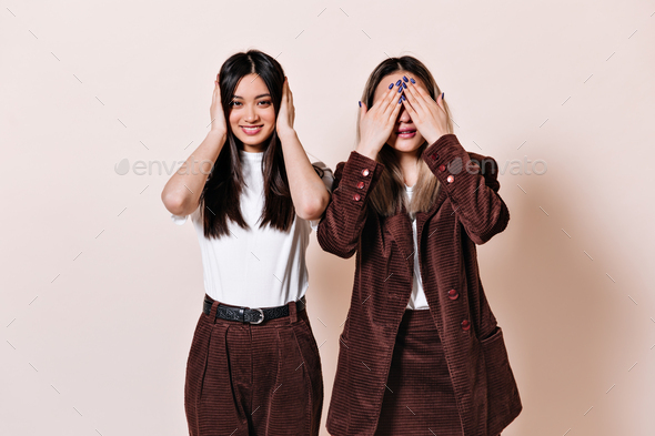 Dark-haired girl smiles and covers her ears, blonde woman in corduroy suit covers her eyes with her