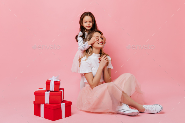 Lady in pink midi skirt and white t-shirt is sitting on pink background while little girl closes he