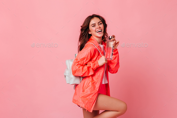 Active slim girl in sports jacket and with backpack laughs and holds bottle of water on pink backgr