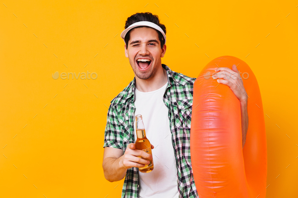 Portrait of brunet guy in white cap pointing to camera. Man with rubber ring holding beer