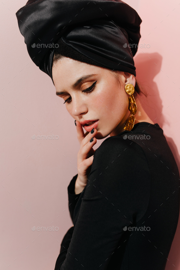 Inspired girl in black turban looking down. Studio shot of well-dressed young woman wears golden ac
