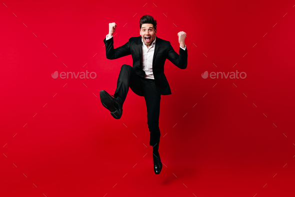 Man in elegant suit fooling around in studio. Emotional male model jumping on red background.