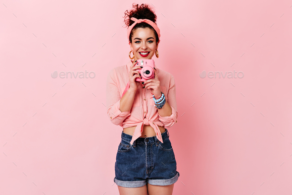 Curly lady in pink blouse and denim shorts is smiling and holding mini camera on isolated backgroun
