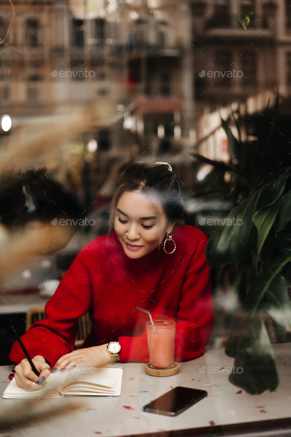 Cute woman dressed in red sweater makes entry in notebook while sitting at table in cozy cafe. Girl