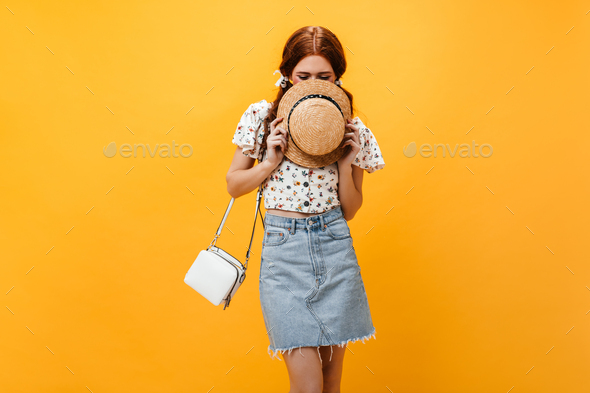 Portrait of mischievous girl covering her face with straw hat. Lady dressed in light denim skirt an