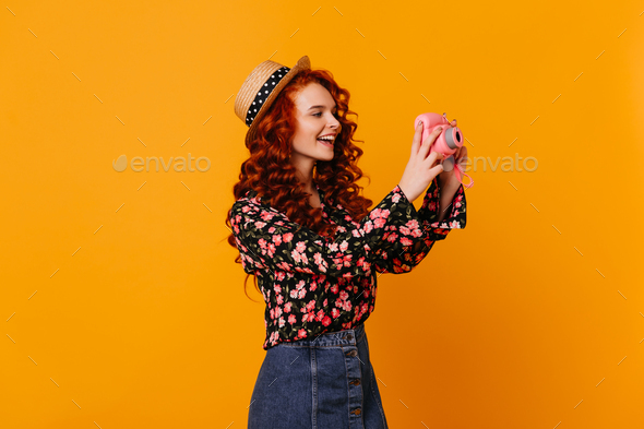 Teen girl in denim skirt and stylish blouse happily makes photo, holding mini camera on orange back