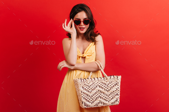 Stylish brunette in yellow sundress puts on glasses in shape of hearts. Girl with beach bag posing