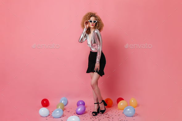 Curly lady in silver blouse and mini skirt wears trendy glasses posing in pink studio among multico