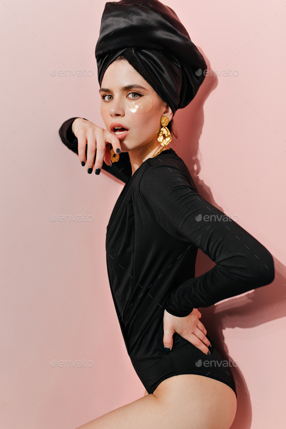 Shapely woman biting finger on pink background. Attractive girl in black turban and bodysuit lookin