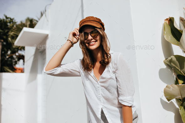 Woman in high spirits coquettishly touches her wavy hair. Lady in cotton shirt and corduroy cap is