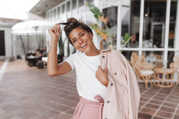 Positive brunette girl in white T-shirt and pink jacket touches her hair band and smiles against ba